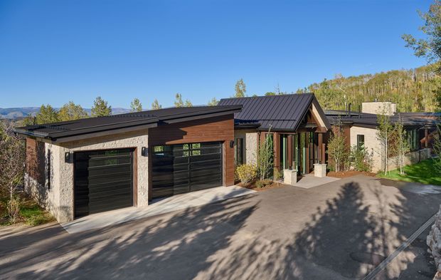 Exterior view of a modern single-storey residence on a sloping, wooded site. The building has low-pitched dark metal roofs with multiple wings, light textured masonry walls, warm timber cladding around the entry, large dark-framed glazing, and integrated double garage doors. A paved driveway and landscaped planting surround the home, with hills visible in the distance.