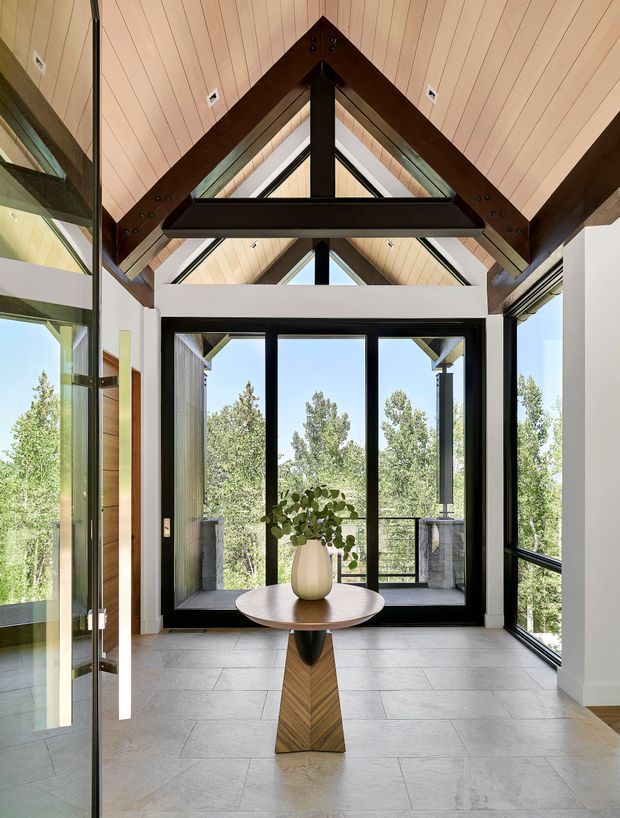 A bright, double-height entry hall with dramatic exposed dark timber trusses and a steeply pitched, light timber-lined ceiling. Full-height black-framed glazing surrounds the space, opening to a balcony and leafy views. The tiled floor and sculptural central console table create a refined, contemporary lodge-like entry.