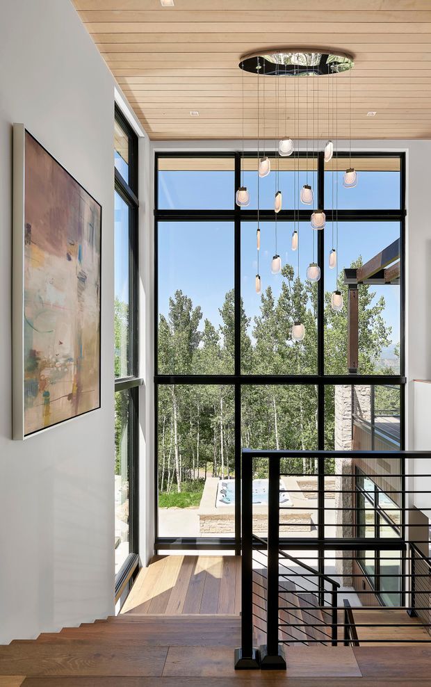 A bright, modern stair landing with a dramatic double-height glazed wall overlooking trees, a pool terrace, and a neighboring outdoor structure. The space features warm timber plank flooring and ceiling lining, black metal-framed floor-to-ceiling windows, a black steel stair balustrade with horizontal rails, and a cluster of suspended pendant lights. A large abstract artwork hangs on the pale wall beside the staircase.