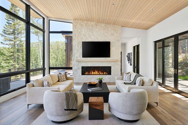 A bright, sophisticated living room with a pair of sofas and rounded lounge chairs arranged around a black coffee table. A large stone fireplace with a linear firebox and wall-mounted television forms the focal point. Floor-to-ceiling black-framed glazing opens views to a wooded landscape, while a warm timber-lined ceiling and pale timber floor create a natural, inviting atmosphere.