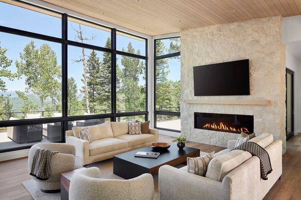 A bright contemporary living room with floor-to-ceiling black-framed glazing overlooking a wooded landscape. The room features a large pale natural-stone fireplace wall with a recessed linear gas fire and mounted television, warm timber ceiling boards, timber flooring, a light neutral sofa and armchairs, and a dark rectangular coffee table.