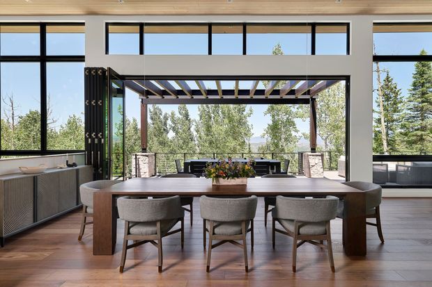 A contemporary dining area with a long wooden dining table and upholstered chairs, positioned beside expansive black-framed glazing. The open wall leads to a covered outdoor entertaining terrace with a dining or lounge setting, stone-clad columns, and views of trees beyond. Warm timber ceiling lining and timber flooring contrast with white walls and dark window frames.