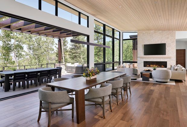 A spacious open-plan dining and living area with a long timber dining table and upholstered chairs in the foreground, opening to an outdoor dining deck through extensive black-framed glazing. The living zone includes a pale stone fireplace with a built-in fire and television, upholstered seating, and a warm timber-lined ceiling. The room has abundant natural light, strong indoor-outdoor connection, and views of surrounding trees.