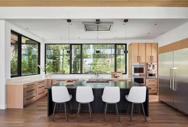 A bright, spacious contemporary kitchen with a large central island and four stools. The room features extensive black-framed glazing overlooking greenery, light timber cabinetry, dark island cabinetry, pale stone benchtops, integrated stainless-steel appliances, timber flooring, and a white ceiling with pendant lights and a roof-mounted skylight or ventilation unit.