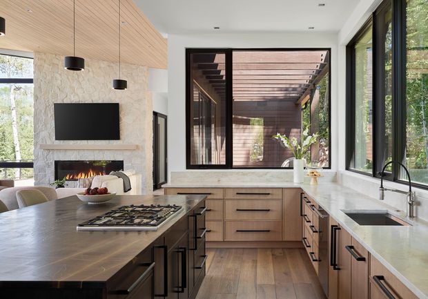 A spacious, contemporary open-plan kitchen with warm timber cabinetry, a large dark island, pale stone benchtops, black-framed windows, and a timber-lined ceiling. A stone fireplace with a television sits beyond the kitchen, while a glazed opening provides views to a timber-clad outdoor room or courtyard. Black pendant lights and hardware add contrast.
