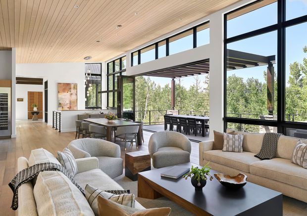 A bright, open-plan contemporary living and dining area with expansive black-framed glazing opening to a covered outdoor entertaining space. The room features a light timber-lined ceiling, pale timber flooring, a neutral seating arrangement around a dark rectangular coffee table, and a dining table positioned beside the windows. A kitchen or service area is partially visible in the background, with abundant views of surrounding greenery through the glazing.