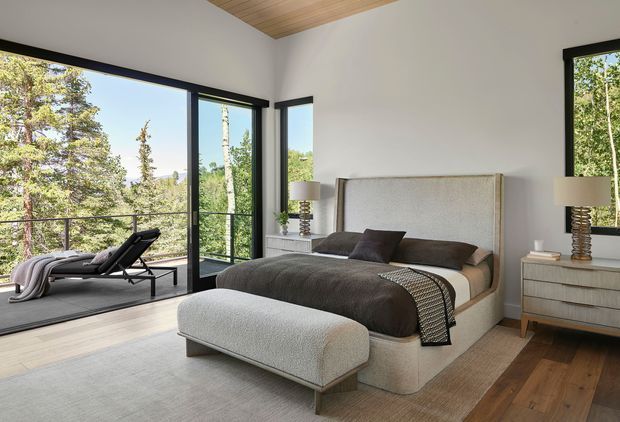 A bright, contemporary bedroom with a large upholstered bed, dark bedding, matching bedside tables and table lamps, and a textured upholstered bench at the foot of the bed. Floor-to-ceiling corner glazing opens to a forested outlook and a small adjoining balcony or terrace with a lounge chair. The room features white walls, a warm timber ceiling, dark window frames, and mixed light and medium timber flooring.