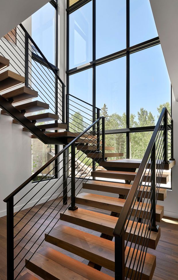 A bright, contemporary interior stairway with open-riser timber treads and dark steel stringers and railings. The multi-level stairwell is framed by expansive floor-to-ceiling glazing, bringing in abundant natural light and views of trees outside. The stair balustrade uses slim horizontal metal cables or rods, while timber flooring and white walls create a clean, warm contrast with the black metalwork.
