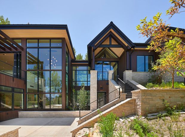 Exterior view of a contemporary multi-level residence on a sloped site, featuring dark timber-toned cladding, extensive floor-to-ceiling glazing, prominent gabled roof forms, exposed timber detailing, and broad stone retaining walls. A landscaped stairway and paved forecourt lead to the entry.