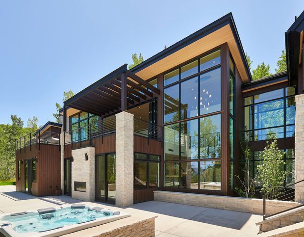 Exterior view of a contemporary multi-level residence with expansive floor-to-ceiling glazing, dark structural framing, warm timber soffits and cladding, pale stone masonry, and a projecting pergola over the terrace. A spa pool occupies the foreground courtyard, while landscaped trees are visible around the building.