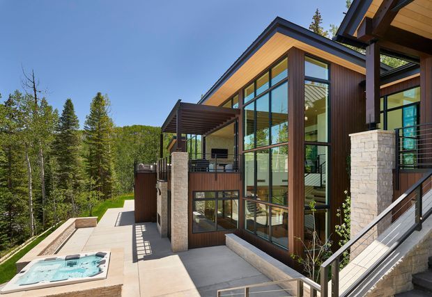 Exterior view of a modern multi-level home set among dense woodland. The house features warm timber cladding, extensive floor-to-ceiling glazing with dark frames, stone-faced columns, projecting roof forms, and elevated terraces. A small outdoor spa or pool is visible at the lower left, with stairs and paved outdoor circulation connecting the levels.
