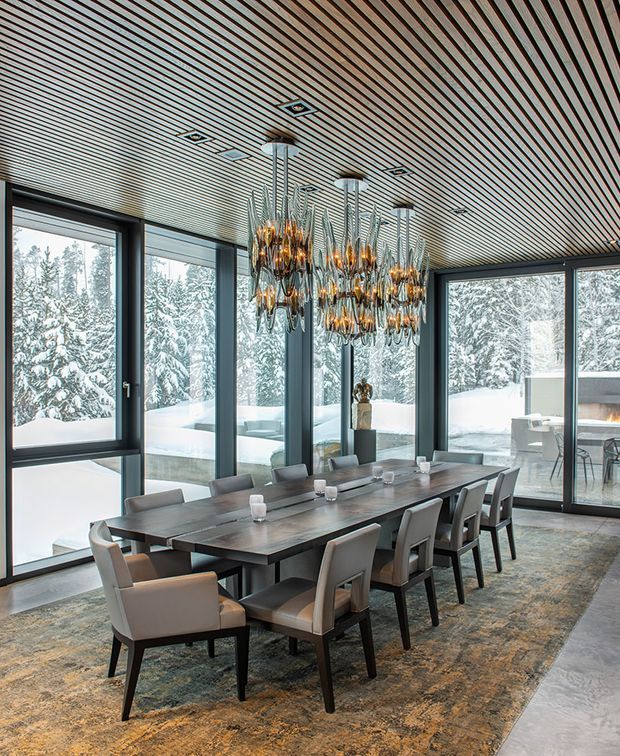 A sophisticated dining room with a long dark timber dining table surrounded by upholstered dining chairs. Floor-to-ceiling glazing frames a snowy forest landscape, while multiple sculptural amber glass pendant lights create a dramatic focal point. The ceiling features linear timber slats, and the room has a polished contemporary alpine-lodge feel.