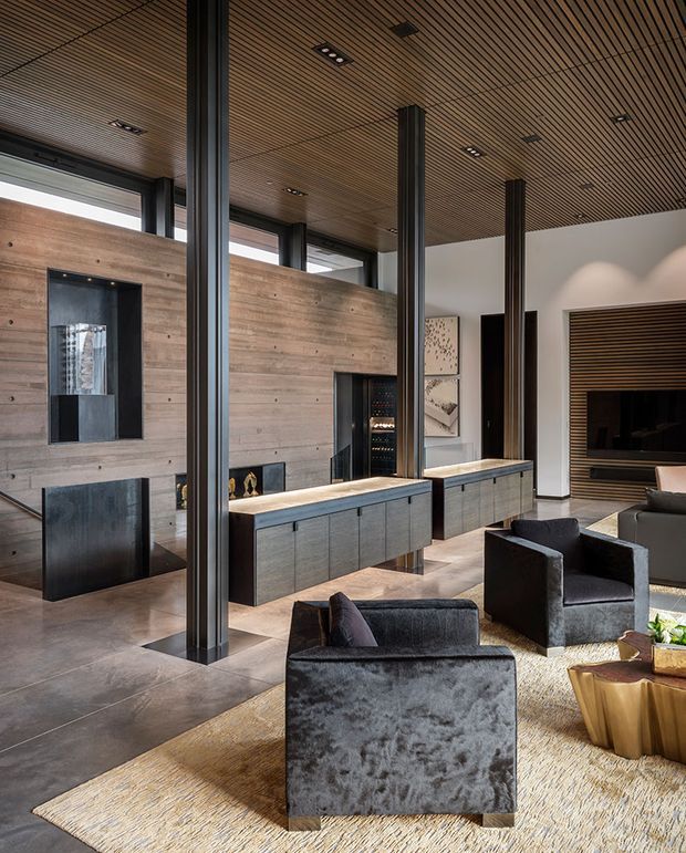 A spacious, refined living room with a dramatic double-height volume, exposed dark structural columns, extensive horizontal timber wall and ceiling lining, and integrated low cabinetry. A linear fireplace, built-in display and wine storage, large artwork, and a dark media wall create a sophisticated entertaining space. Plush charcoal armchairs and sofas sit on a large neutral rug over a polished stone or concrete floor.