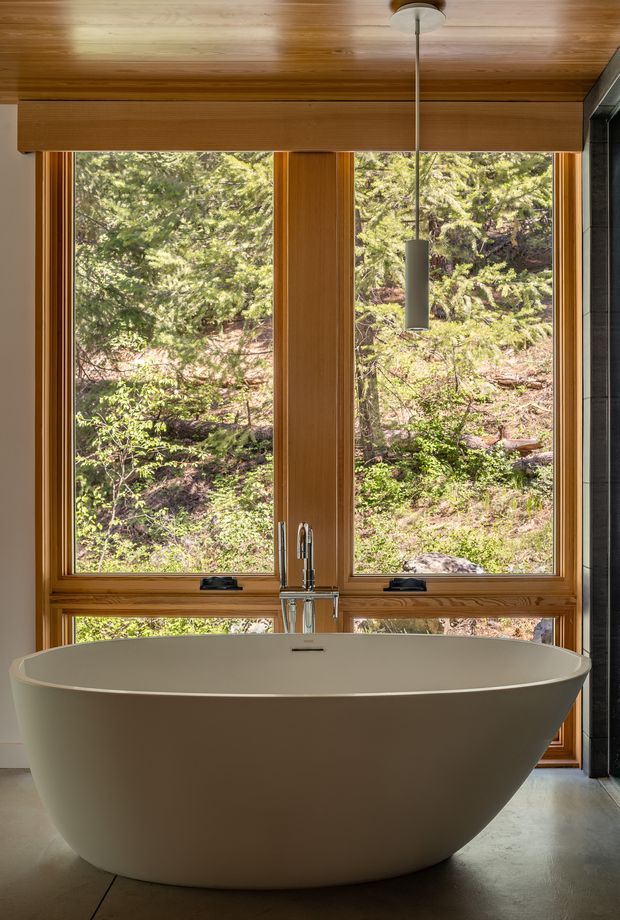 A serene bathroom centered on a large freestanding oval bathtub positioned in front of expansive timber-framed windows overlooking dense woodland. Warm timber surrounds the glazing and continues across the ceiling, with a slim cylindrical pendant light above the tub, polished chrome tapware, and a dark vertical wall surface at the right edge. The space has a calm, nature-connected contemporary design.