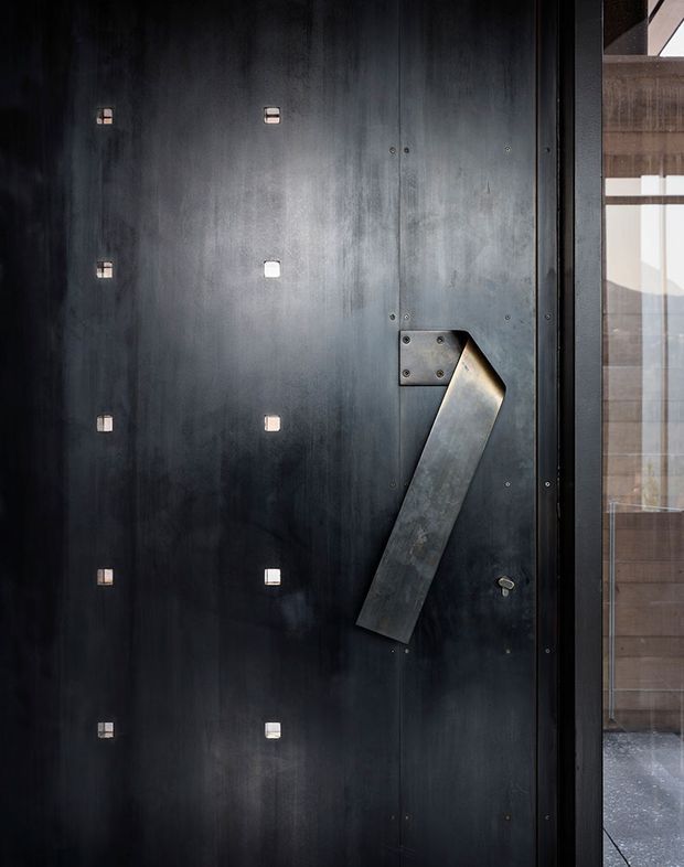 Close-up of a dark, industrial-style entry door with a broad weathered metal surface, exposed fasteners, small square inset details, and a large angular metal pull handle. A dark metal-framed glazed panel or sidelighting is visible at the right, with part of the building beyond.