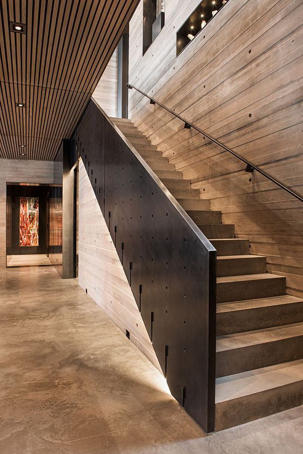 A dramatic contemporary interior stairway with exposed concrete and warm timber finishes. The staircase has broad concrete treads, a dark perforated metal side balustrade, and a slim wall-mounted handrail. Horizontal timber wall cladding and a slatted timber ceiling create strong linear detailing, while recessed and concealed lighting accentuates the stair and double-height volume. A polished concrete floor and a lit artwork are visible in the hallway beyond.