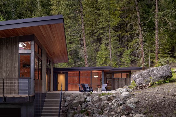 A modern single-storey cabin-style residence set among dense forest and exposed granite rocks. The building has dark vertical timber cladding, expansive timber-framed glazing, warm timber soffits, and low-pitched dark metal roofs. A covered outdoor terrace with lounge chairs connects the separate-looking wings, while steps and a rocky landscape lead up to the entry.