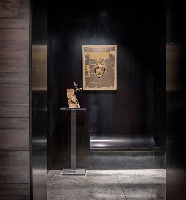 A dark, dramatic display niche with black reflective wall panelling, a framed artwork and a small sculptural object presented on a slender pedestal. The niche is framed by dark timber or metal trim, with a grey polished stone or concrete floor and focused accent lighting.