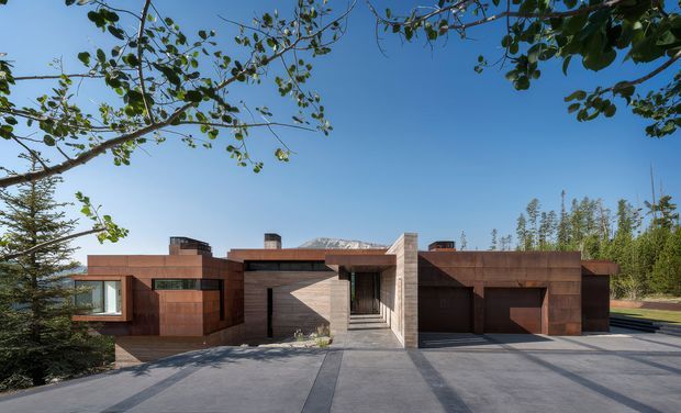 A low, single-storey contemporary house with a strongly horizontal composition, clad in warm rust-brown panels and masonry. Deep roof overhangs, recessed entry, a projecting glazed volume, and a large integrated garage face a broad pale concrete driveway. Mature trees frame the foreground, with a clear blue sky and distant mountain ridge visible behind the home.