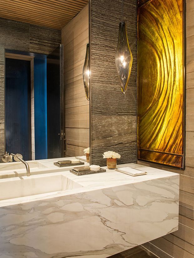 A luxurious contemporary bathroom featuring a large freestanding white marble bathtub with integrated marble surround, textured dark stone wall panels, warm horizontal timber ceiling and wall lining, sculptural pendant lights, and a dramatic backlit amber feature panel. A dark-framed doorway or shower enclosure is visible in the background.