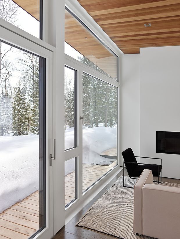 A contemporary living room with expansive floor-to-ceiling glazing and a full-height glazed door overlooking a snow-covered wooded landscape. The room features a warm timber-lined ceiling, white walls, pale framed windows and doors, a wall-mounted dark fireplace, a black lounge chair, a light upholstered sofa, a woven rug, and timber decking visible outside.