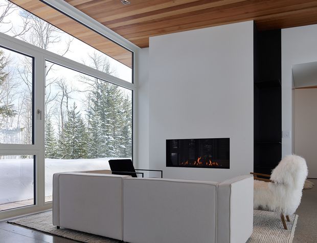 A bright contemporary living room with floor-to-ceiling glazing overlooking a snowy forested landscape. The room features a large pale sectional sofa, a minimalist white fireplace wall with a linear firebox, dark built-in shelving, a timber-lined ceiling, and a white furry accent chair on a textured rug.
