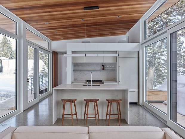 Bright modern kitchen in a glazed living space, featuring a central white island with integrated cooktop and sink, three timber bar stools, white cabinetry, a marble-look splashback, polished concrete-style flooring, and extensive floor-to-ceiling windows. The ceiling is clad in warm timber boards, with snowy trees visible outside.