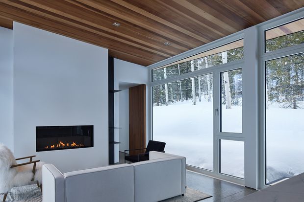 A contemporary living room with a linear built-in gas fireplace set into a white wall, integrated dark timber shelving and cabinetry, a pale grey sectional sofa, and a black lounge chair. Floor-to-ceiling glazing and a glazed door open views to a snowy wooded landscape. The room features a striking warm timber slatted ceiling and cool grey window frames, creating a refined, modern alpine atmosphere.