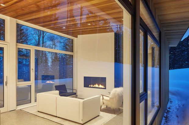 A refined contemporary living room viewed through extensive glazing, with a white sectional sofa arranged around a linear built-in fireplace. The room features warm timber-clad ceiling boards, pale walls, large framed windows and doors, and a light neutral floor. Snow-covered landscaping and trees are visible outside at dusk, while reflections in the glazing add depth to the scene.