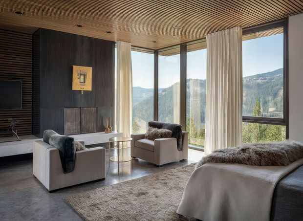 A sophisticated living room with floor-to-ceiling corner glazing overlooking a mountainous landscape. The room features a pair of upholstered armchairs, a low glass side table, a large textured rug, and a partially visible sofa or daybed layered with neutral throws and fur-textured cushions. Dark timber wall panelling and a linear timber slat ceiling contrast with pale upholstery, while full-height curtains frame the expansive view.
