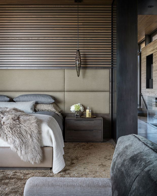 A sophisticated bedroom interior with a large upholstered bed, layered neutral bedding, a shaggy throw, and a dark wood bedside cabinet. The feature wall combines horizontal timber battens with a padded beige upholstered headboard. A sculptural pendant hangs beside the bed, while a dark structural column frames a view toward an adjacent timber-lined area. The palette is warm, muted, and textural, with contemporary detailing and soft natural materials.