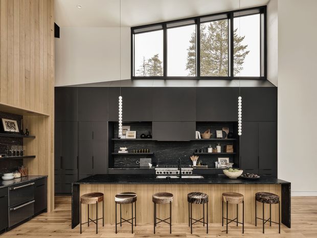 Contemporary kitchen with a large dark charcoal built-in cabinetry wall, open display shelving, black stone island benchtop, and a timber-clad island front. Six upholstered bar stools line the island. Tall clerestory-style black-framed windows bring in natural light, while light timber wall panelling and flooring add warmth. Decorative pendant lights hang over the island.