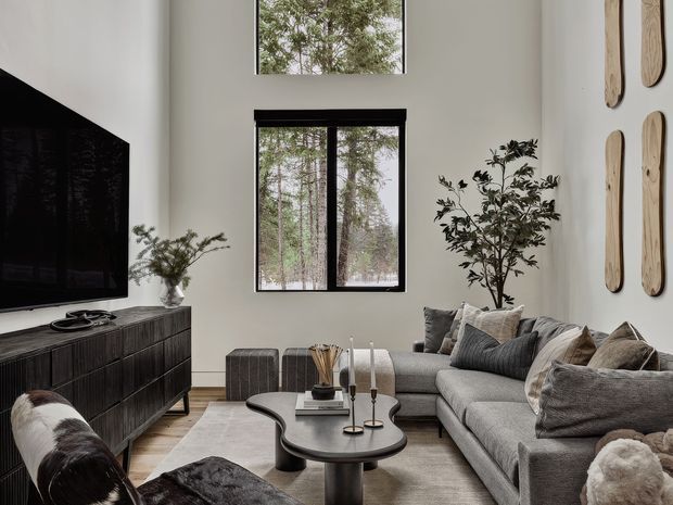 A refined contemporary living room with a tall light-coloured wall, double-height glazing, and a large black-framed window overlooking trees. The room features a charcoal sectional sofa with layered neutral cushions, a pair of round black coffee tables, a dark fluted media console with a wall-mounted television, textured neutral rug, sculptural wall pieces, and indoor plants. The palette is predominantly warm grey, beige, black, and off-white, creating a calm, sophisticated atmosphere.