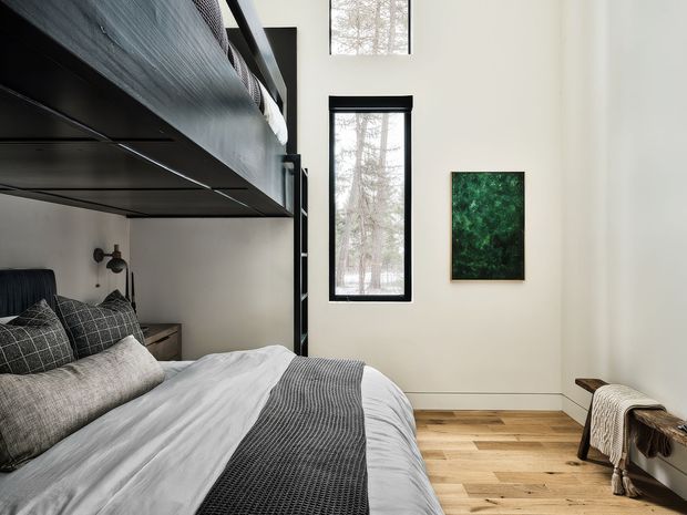 A contemporary bedroom with a lofted sleeping platform and integrated black structure along the left side. The room features a neatly made bed with layered grey and white bedding, dark textured cushions, a minimalist bedside table and wall-mounted reading lights. Tall narrow black-framed windows bring in views of trees, while pale walls, light timber flooring, and a green-toned artwork create a calm, refined interior. A simple wooden bench with a draped throw sits along the right wall.