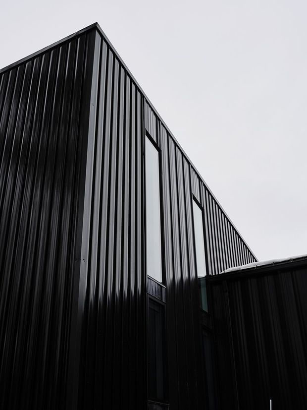 Upward-looking architectural exterior view of a sharply angular, gable-like building form clad in dark vertically ribbed metal panels. Tall narrow windows punctuate the facade, while a lower adjoining volume projects across the foreground. The monochrome black and grey composition emphasizes the corrugated texture, geometric roofline, and contemporary industrial character.