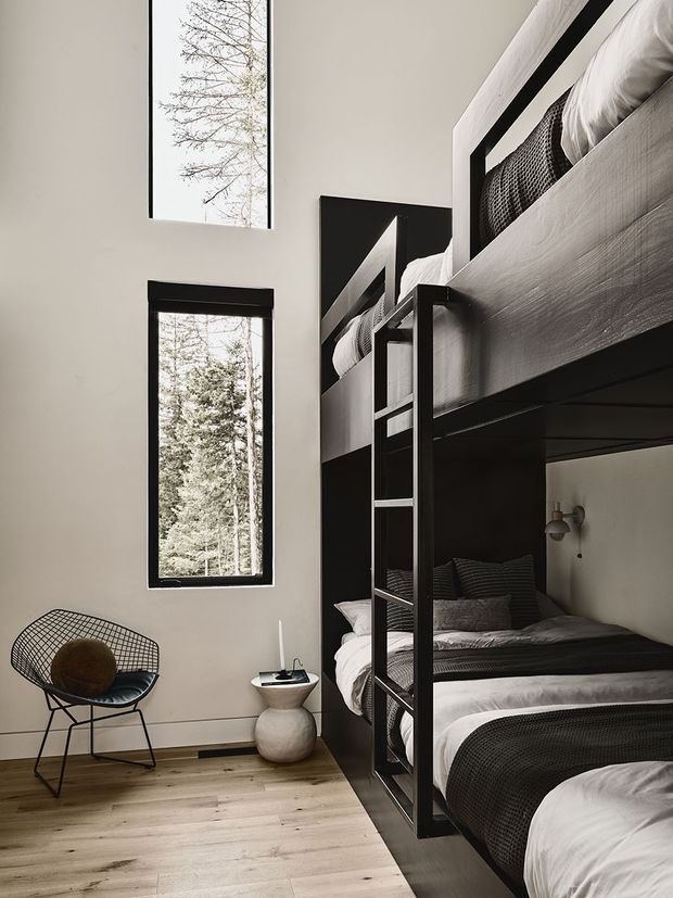 A tall, narrow bedroom with built-in black bunk beds arranged along one wall. The beds feature light wood-grain panels, dark bedding, and integrated ladder shelving. Pale walls, warm timber flooring, slim black-framed vertical windows, and a sculptural wire lounge chair create a restrained, contemporary interior. A small rounded bedside table and wall-mounted light sit beside the lower bed.