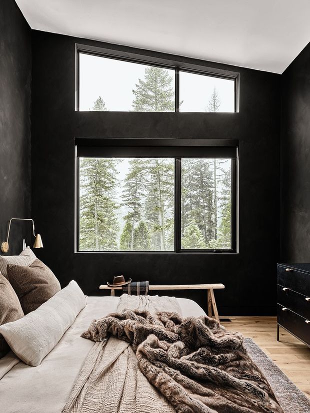 A moody, contemporary bedroom with near-black textured walls, a white ceiling, and large black-framed windows overlooking a forest. The room features a low upholstered bed layered with neutral bedding and a textured fur throw, a slim timber bench at the foot of the bed, a minimalist floor lamp, and a dark chest of drawers. Light timber flooring and abundant daylight contrast with the dark wall finish.
