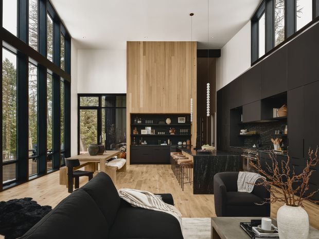 A sophisticated open-plan living, dining and kitchen interior with floor-to-ceiling black-framed glazing, pale timber flooring, a dramatic black built-in kitchen wall and island, and a tall vertical timber feature wall with integrated shelving. Black sofas, a timber dining table, and restrained neutral furnishings create a refined contemporary atmosphere. The glazing brings in views of trees and abundant natural light.