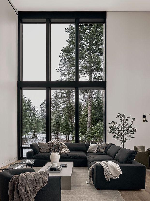 A tall, double-height living room with expansive black-framed floor-to-ceiling glazing overlooking a wooded landscape. The space features a large dark charcoal sectional sofa, neutral textured throws and cushions, a square concrete or stone coffee table, a pale rug, minimalist white walls, and a small indoor tree. The overall design is restrained, contemporary, and natural, with strong vertical window proportions and abundant daylight.