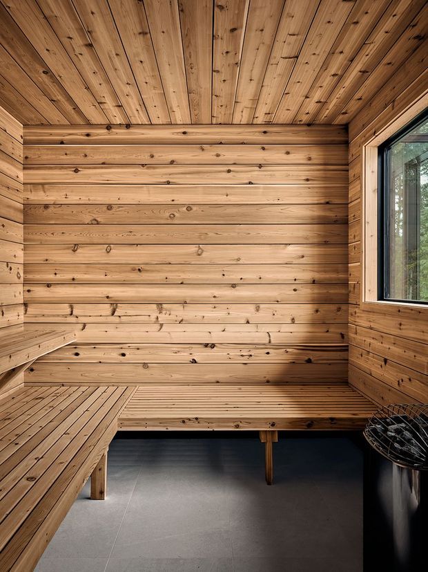 A compact timber-lined sauna interior with horizontal cedar wall boards, a matching slatted ceiling, and built-in tiered benches along the walls. A large framed window brings in views of trees and natural light. The floor is finished in large dark grey tiles, and a black sauna heater is partially visible in the foreground.