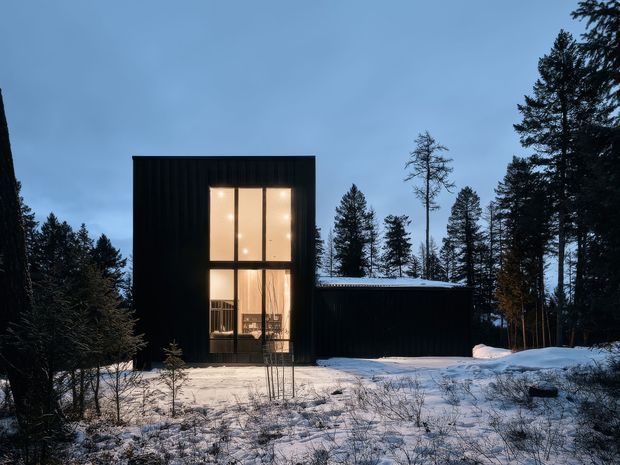 A compact, dark-clad modern house set within a snow-covered forest at dusk. The rectilinear two-storey volume has a tall floor-to-ceiling glazed opening glowing warmly from within, connected to a lower single-storey wing with a low roof. Coniferous trees surround the building, creating a secluded alpine woodland setting.