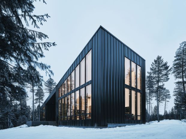 Exterior view of a contemporary dark-clad, gable-roofed building set within a snowy forest. The facade features vertically ribbed black metal cladding and extensive floor-to-ceiling glazing, with warm interior light and reflections of surrounding trees visible through the windows.