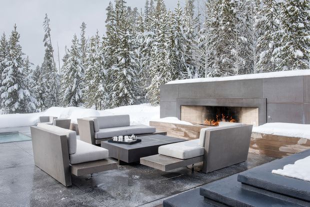 A contemporary outdoor lounge area set in a snowy forest, with low modular grey seating arranged around a dark rectangular coffee table. A large built-in concrete or stone fireplace forms the backdrop, with a fire burning in the open hearth. Snow-covered trees and paving surround the terrace, creating a dramatic winter setting.