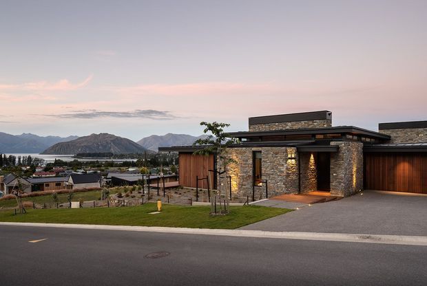 Contemporary single-storey residence viewed from the street at dusk, with a low flat-roofed form, dark timber-clad sections, and prominent rough stone wall cladding. Warm integrated lighting highlights the entry and stone facade. A driveway and garage occupy the right side, while a landscaped front lawn and neighbouring homes lead toward a dramatic mountain and lakeside landscape in the distance.