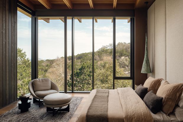 Warm, timber-lined bedroom with a low-profile upholstered bed, layered neutral bedding and cushions, a rounded lounge chair with ottoman, and dramatic floor-to-ceiling glazing overlooking dense vegetation. Exposed timber ceiling beams and dark-framed windows create a refined natural retreat.