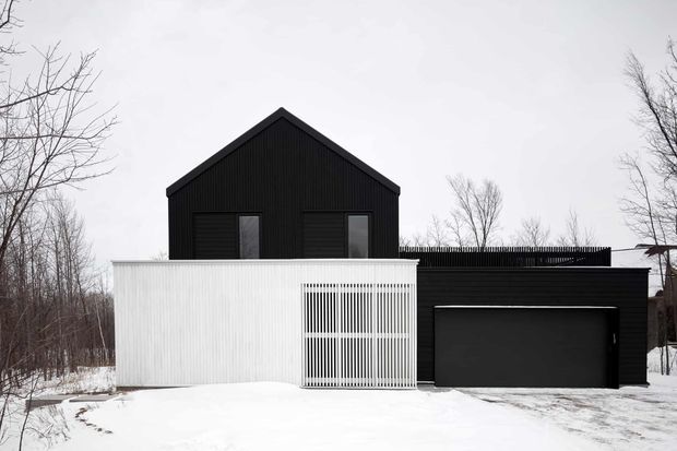 Front exterior view of a compact modern house in a snowy, wooded setting. The building has a simple black gable-roofed upper volume with dark vertical cladding, a white vertically ribbed lower facade and privacy screen, and an attached single-storey black garage volume with a broad dark garage door. Bare deciduous trees and snow surround the property.