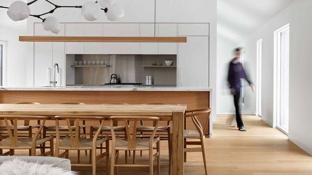 Bright open-plan kitchen and dining area with a long timber dining table and matching wooden chairs in the foreground. The kitchen has white flat-panel cabinetry, a pale stone benchtop, timber overhead shelving, and a stainless-steel sink and tap. A sculptural branching pendant light hangs above the dining zone, while tall windows and light timber flooring create an airy, contemporary interior. A blurred person is visible in the adjoining circulation area.