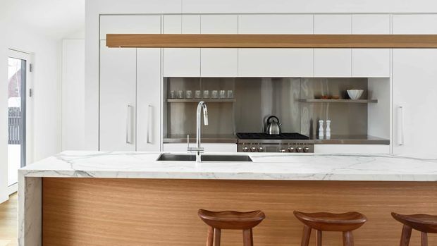 Bright contemporary kitchen featuring a large white marble-look island with integrated sink and chrome tap, warm timber-clad island seating, white full-height cabinetry, and a stainless-steel cooking and display niche with open shelving. Three timber bar stools sit at the island, while a long timber overhead feature adds warmth and definition.