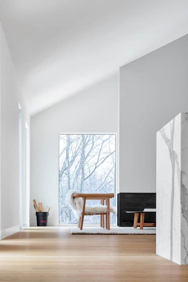 Bright minimalist living room with white walls and a high sloping ceiling, pale timber flooring, and a large window overlooking leafless trees. A simple wooden lounge chair with a sheepskin throw sits beside a low black media cabinet, while a white-and-grey veined marble-clad fireplace or partition anchors the right side of the room. The composition is calm, airy, and sparsely furnished.