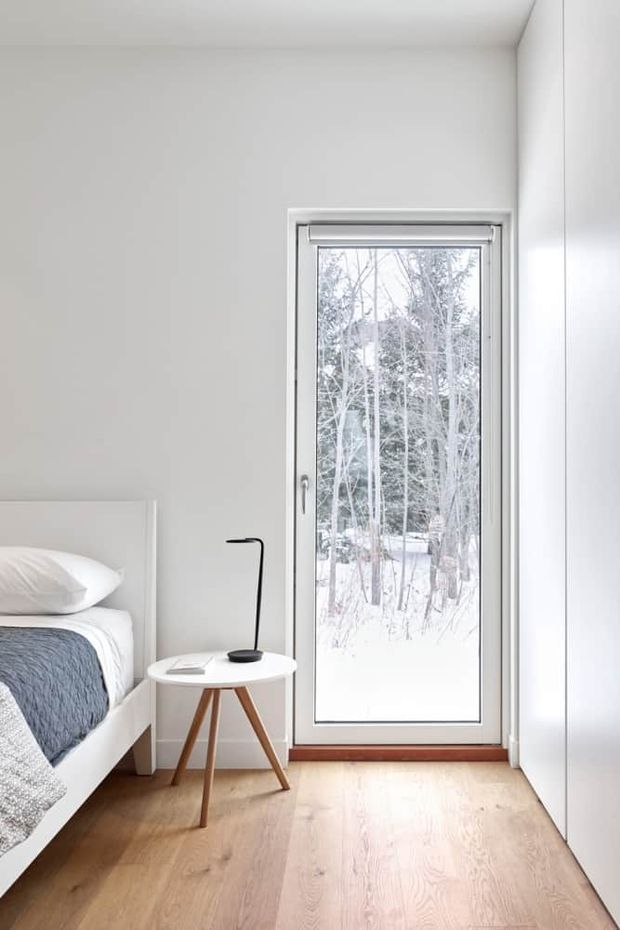 Bright, minimalist bedroom corner with a white bed partially visible at left, white bedding and a textured blue-grey throw, a small round white side table with a slim black lamp, pale timber flooring, white walls and built-in white cabinetry. A full-height glazed door or window opens the view toward a snowy wooded landscape outside.